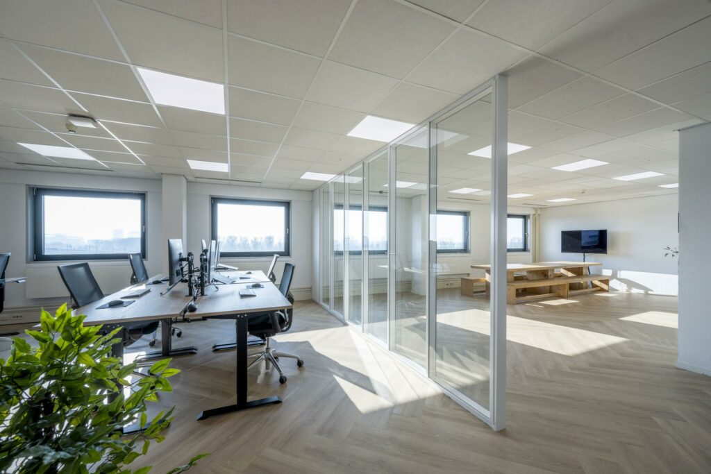 Modern en licht kantoorinterieur aan de Joan Muyskenweg met open werkplekken, glazen vergaderruimte en houten vergadertafel.